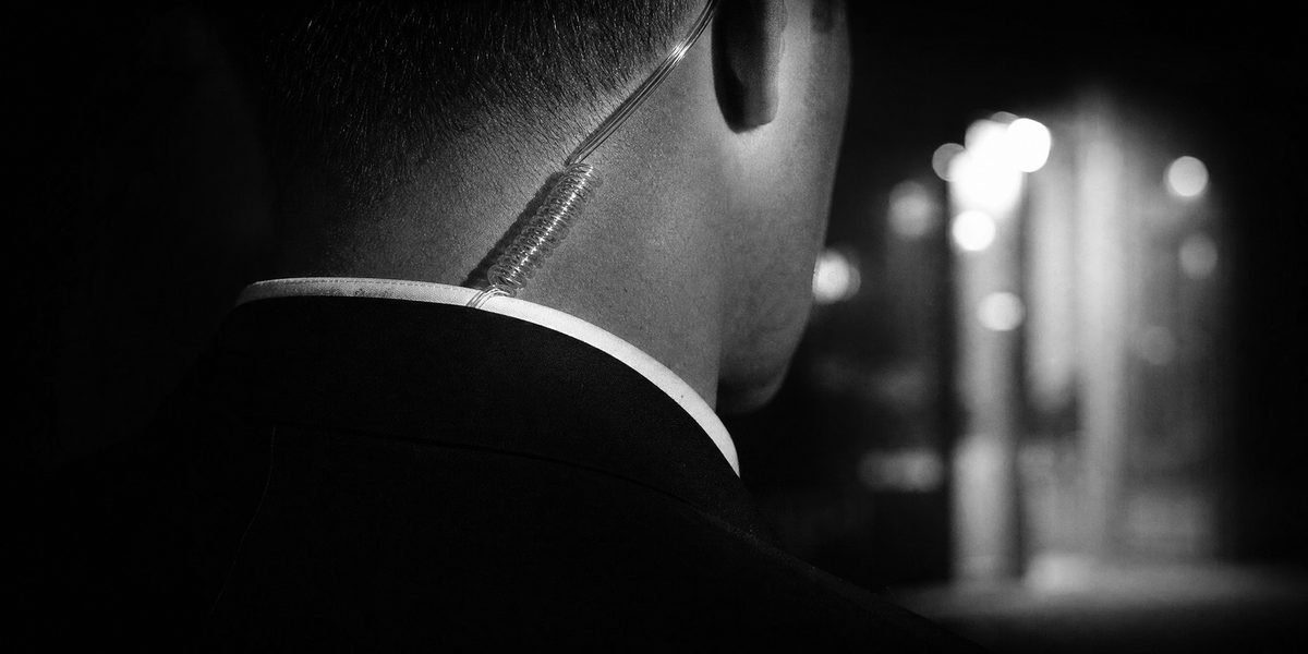 Close protection officer escorting a client in an evening gown and gold clutch outside a hotel at night
