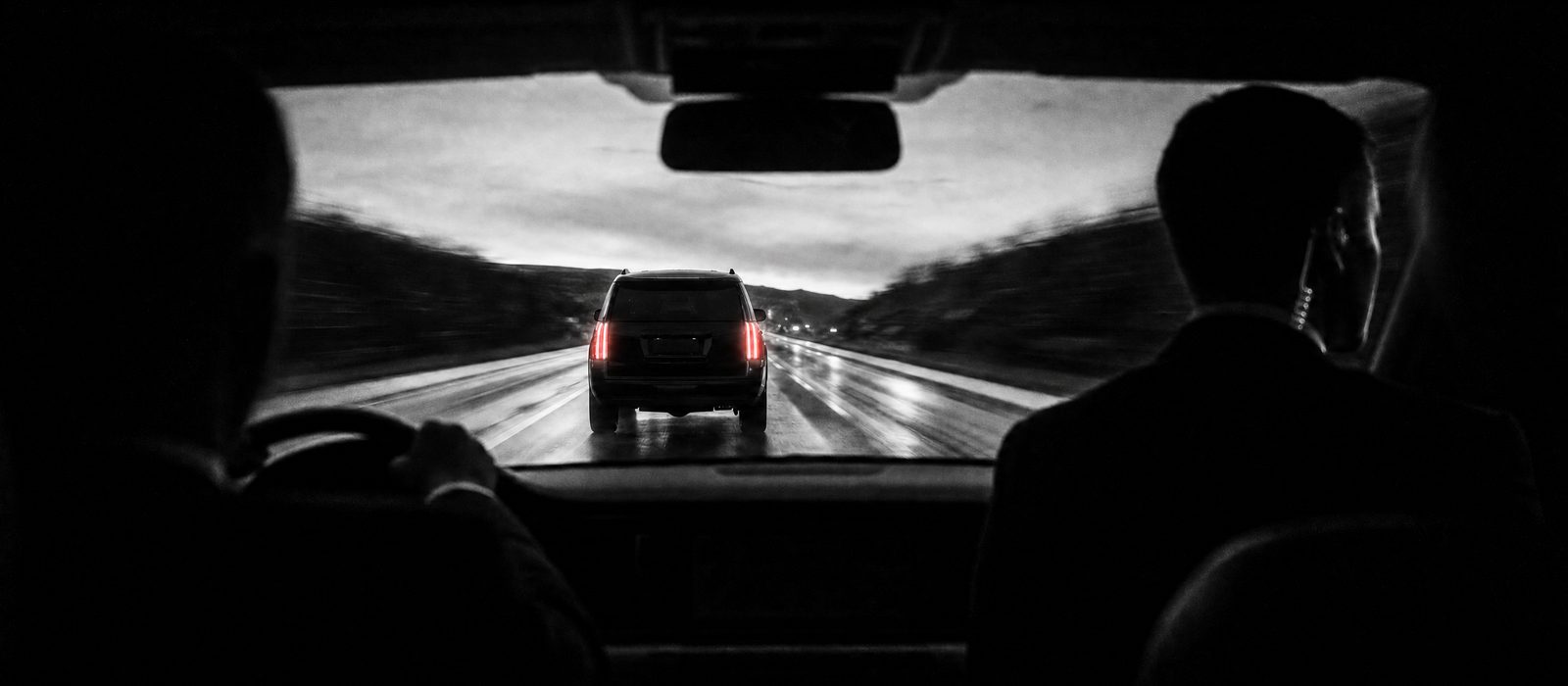 Alacakaranlıkta ıslak yolda güvenlik konvoyu, takip aracından görülen zırhlı SUV