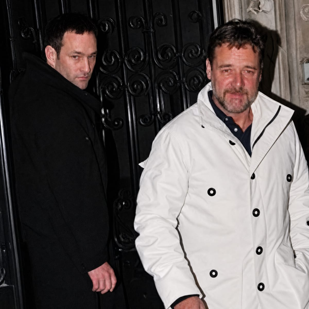Russell Crowe, beyaz paltosuyla süslü bir kapının önünde Agun Security ile